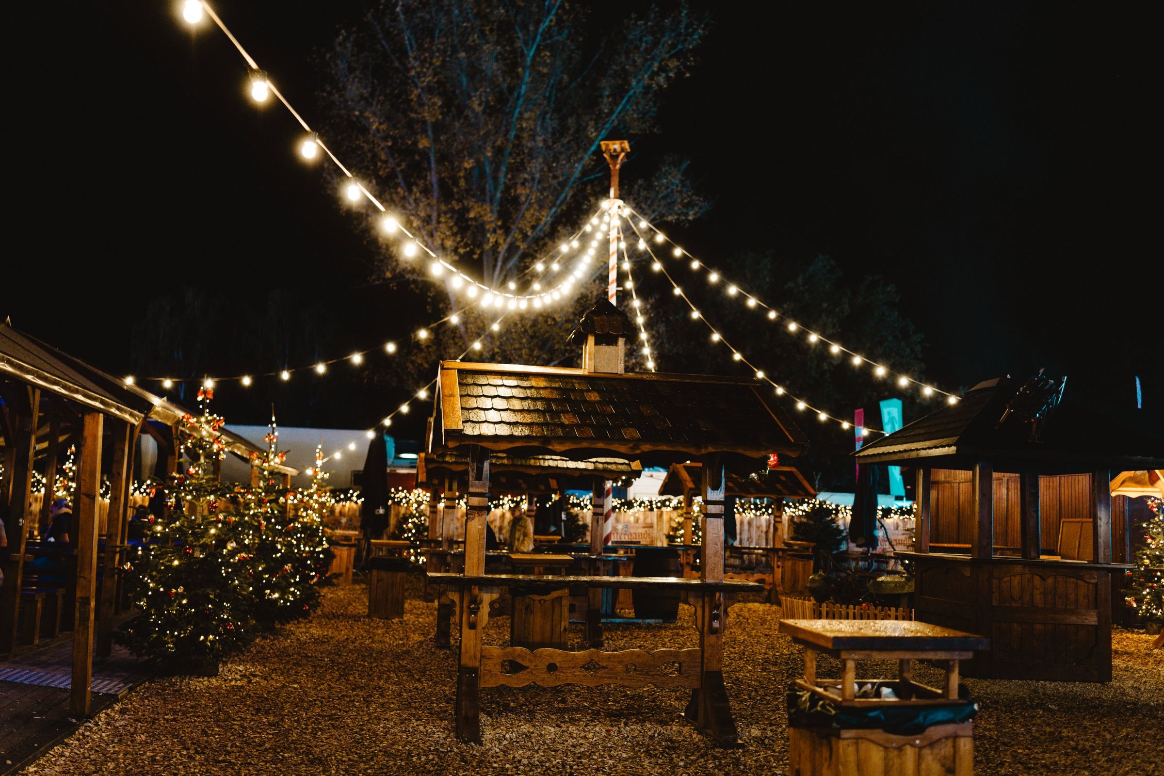 Holzhütten, Tannen und Lichterketten auf einem Weihnachtsmarkt im Wald bei Nacht